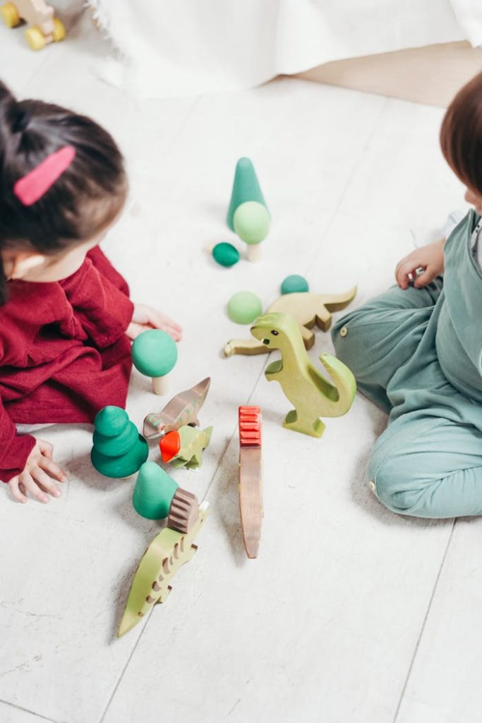 Two children engaged in imaginative play with wooden dinosaur toys on the playroom floor.